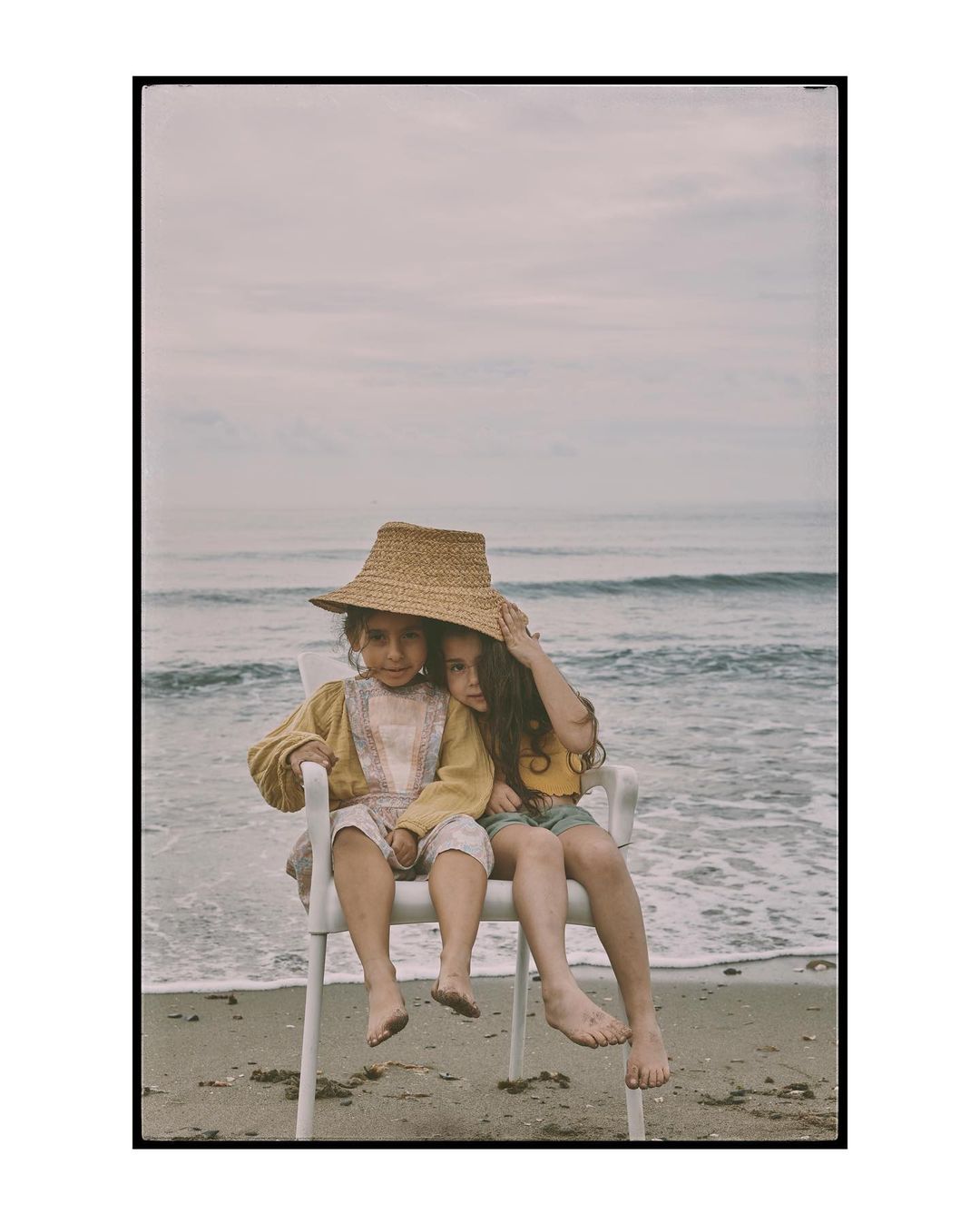 Two young children sit together on a white plastic chair on a sandy beach, wearing casual clothing and a straw hat, with ocean backdrop.