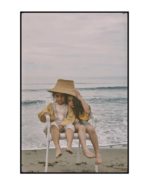 Two young children sit together on a white plastic chair on a sandy beach, wearing casual clothing and a straw hat, with ocean backdrop.