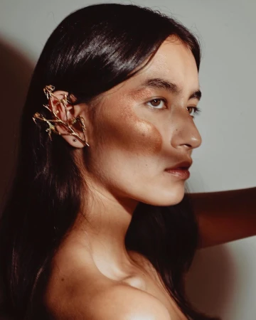 Profile portrait of a woman wearing an intricate gold branch ear cuff and warm metallic makeup by Ian Boddy.