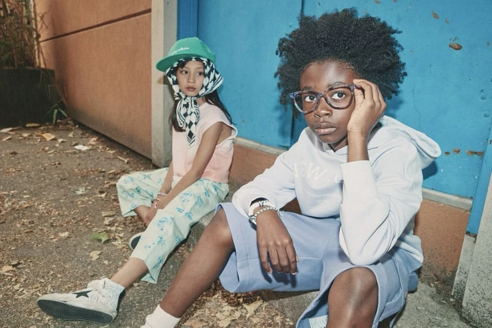 Two stylish children, a young boy and girl, sit together outdoors. The boy wears glasses and a hoodie, the girl a hat and patterned scarf.