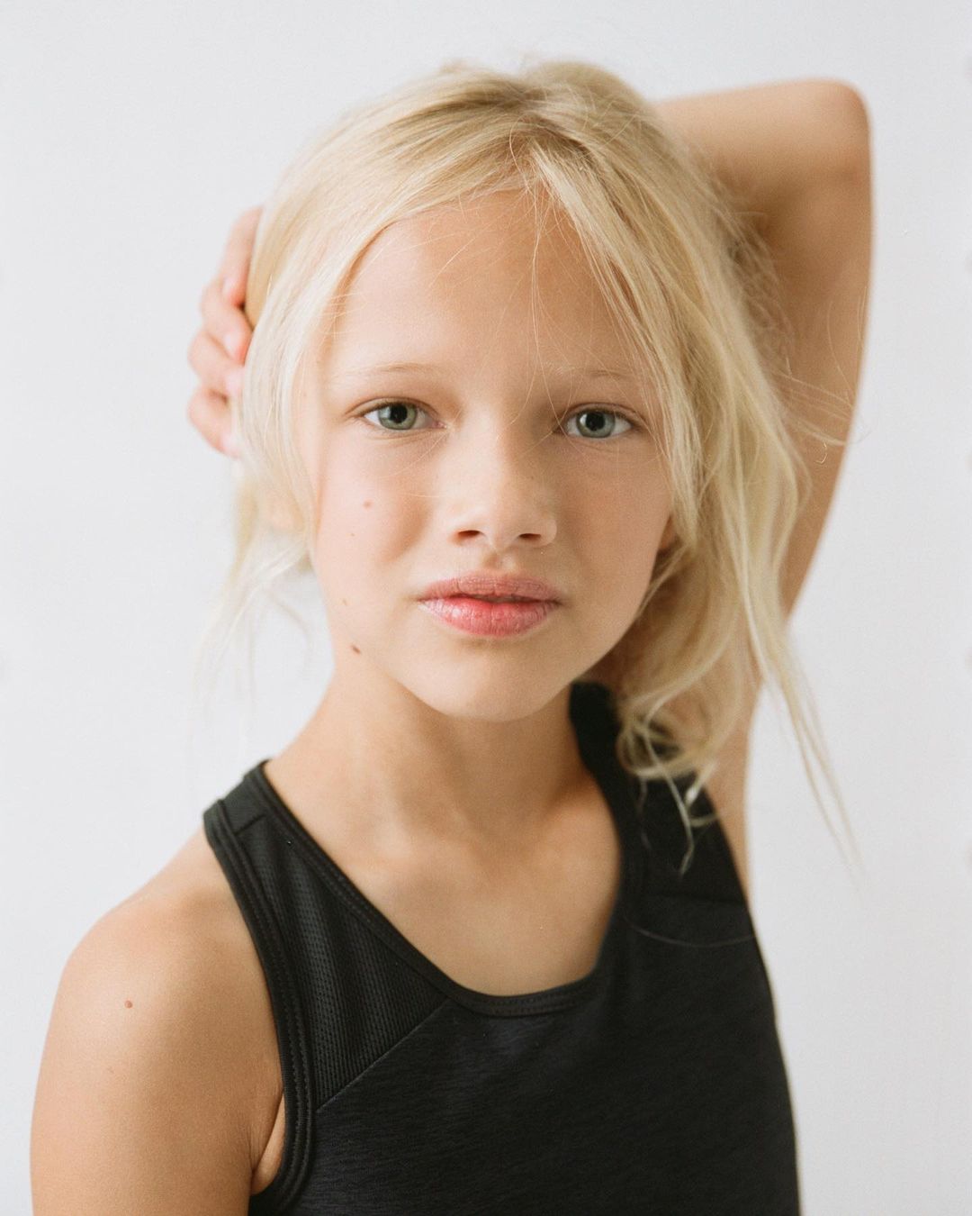 Portrait of a young girl with blonde hair and a black tank top against a white background, looking directly at the camera with a neutral expression.