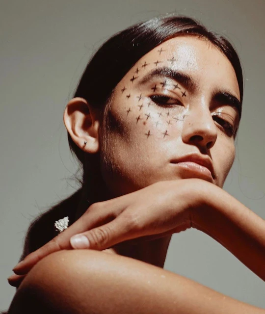 Close-up portrait of a young woman with dark hair and neutral skin tone, wearing a floral earring and black cross face paint design.