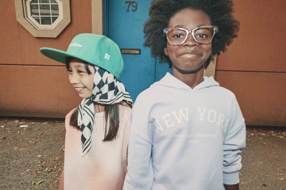 Two smiling children, one wearing a green cap and checkered scarf, and the other wearing glasses and a light purple hoodie, stand outdoors.