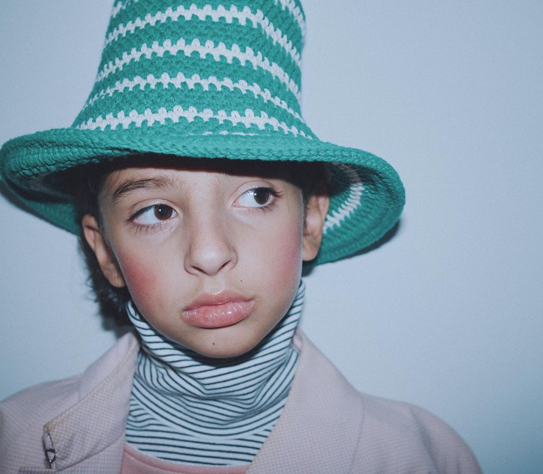Close-up portrait of a child with fair skin wearing a striped green and white bucket hat, pink blush, and a striped turtleneck and pink blazer.