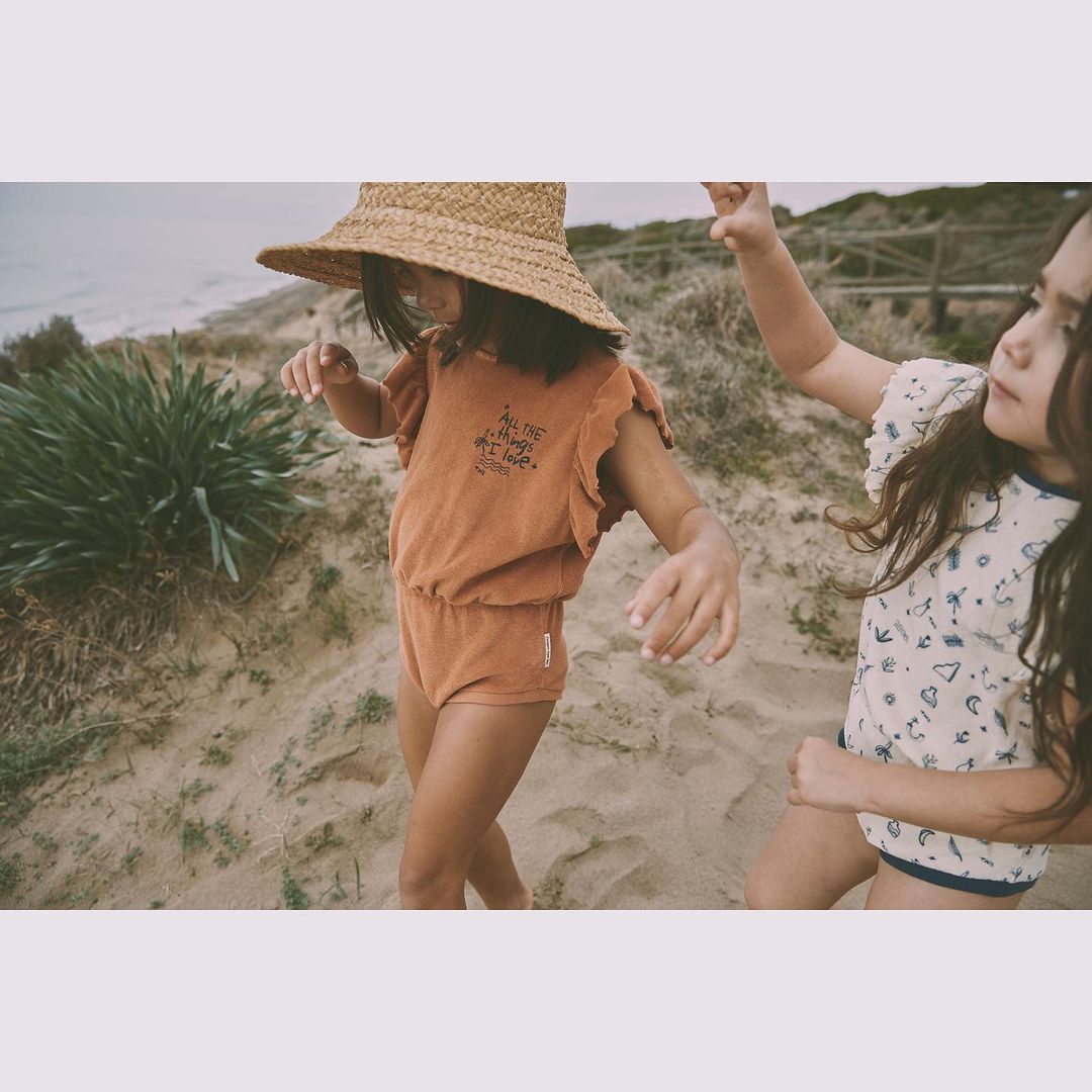 Two young girls on a sandy beach wearing summer outfits and straw hats, enjoying a sunny day by the ocean with a cloudy sky.