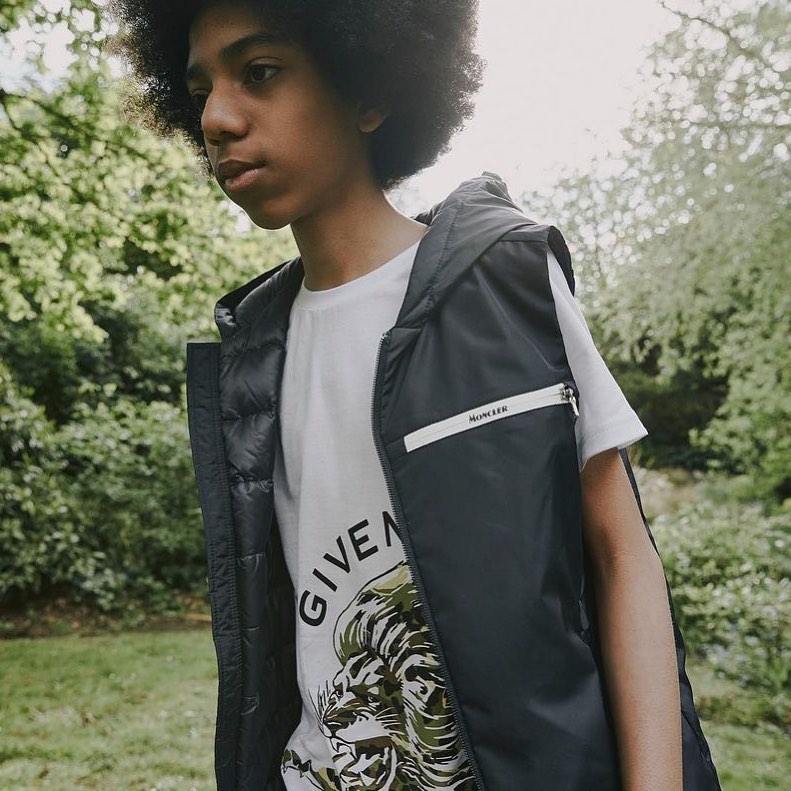 Youth poses in a black vest and Givenchy t-shirt outdoors, trees in background. Fashion shot with shallow depth of field.