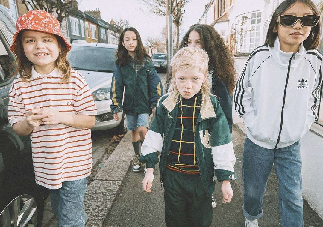 Group of five children wearing vintage clothing on a city street, one in a bucket hat and striped shirt looking at the camera.