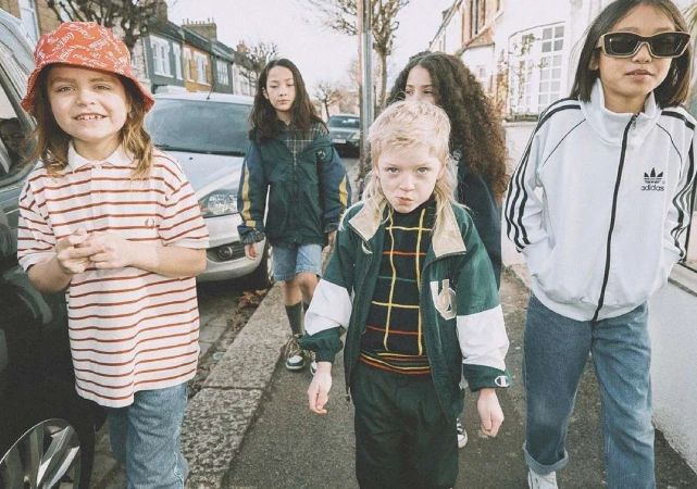 Group of five children wearing vintage clothing on a city street, one in a bucket hat and striped shirt looking at the camera.