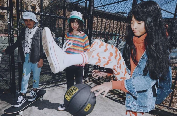 Three children on an urban basketball court. One child kicks a basketball in the foreground with another on a skateboard behind her.