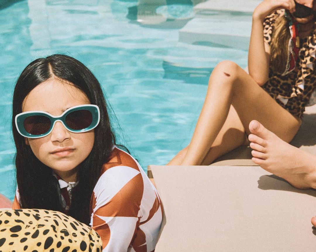 Young girl wearing sunglasses near a pool with an adult in a leopard print swimsuit relaxing in the background on a sunny day.