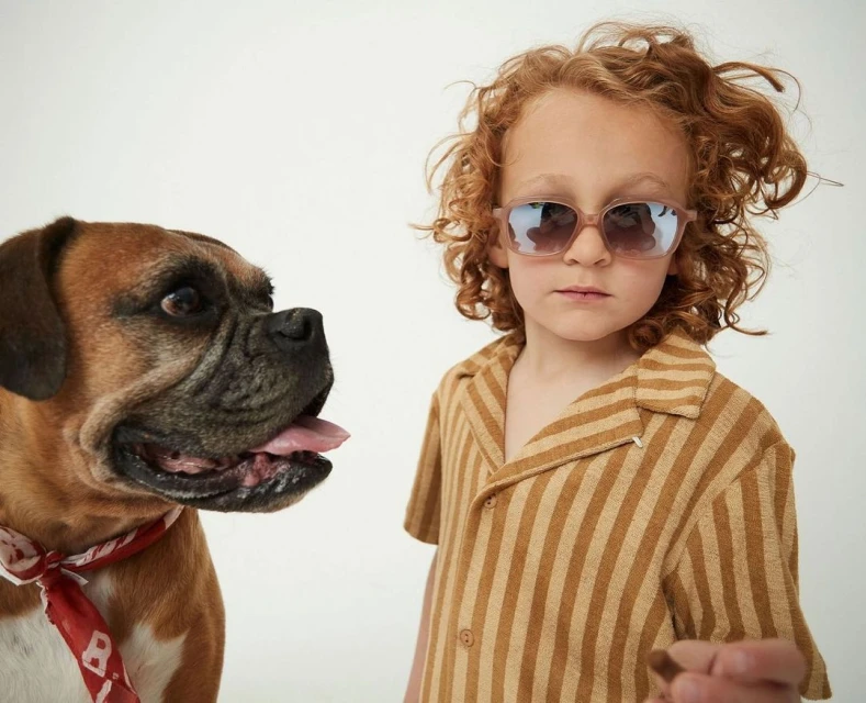 Child with red hair poses with a boxer dog against a white background, showcasing eyewear and casual fashion in a portrait shot.