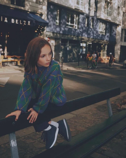 Young girl in a green and blue argyle sweater sits on a bench in a city street on a sunny day, posing for an outdoor fashion shot.