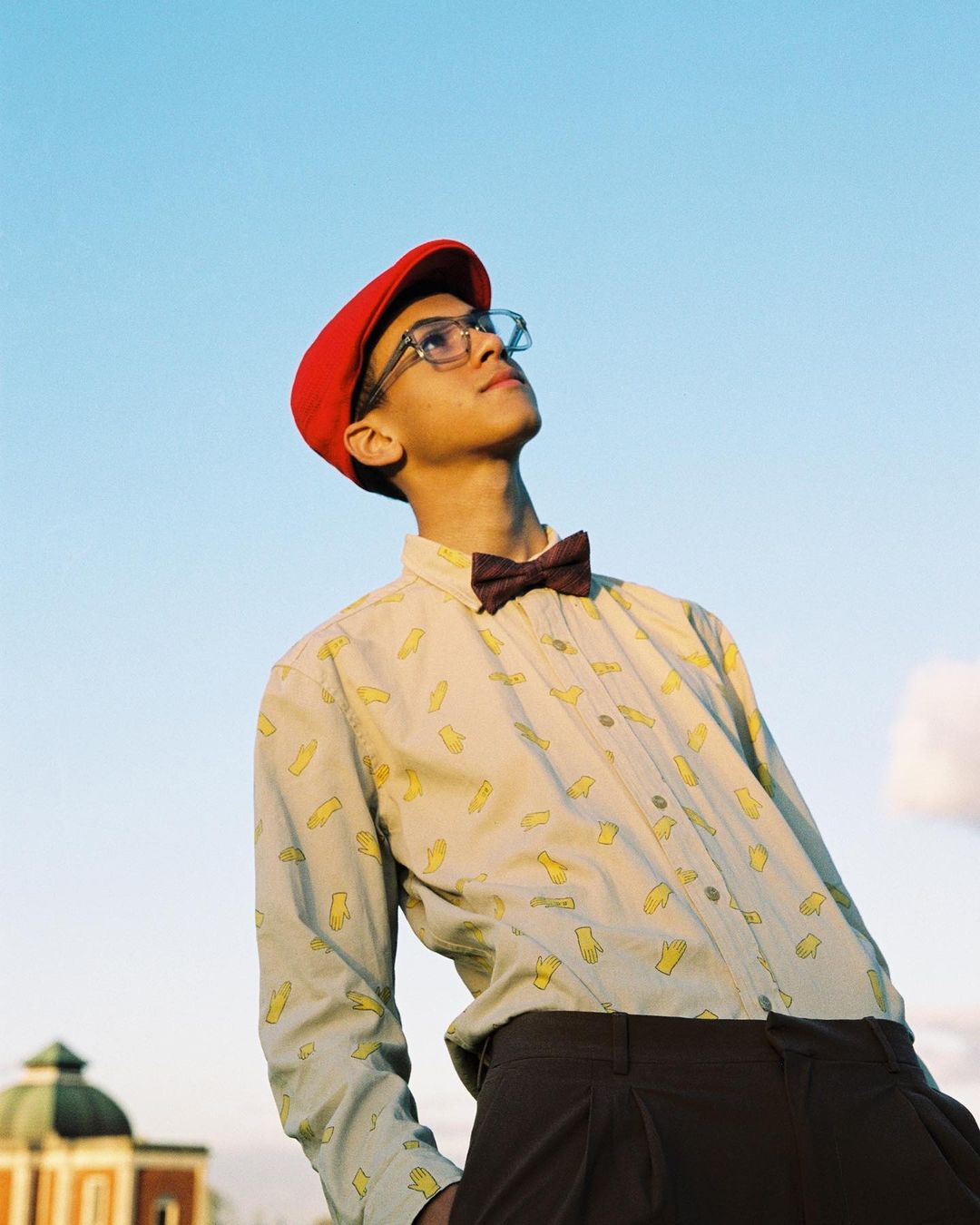 Young man in vintage-inspired outfit gazes upward outdoors against a clear blue sky with a building visible in the background.