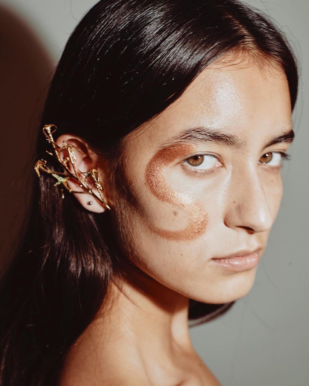 Close-up shot of a woman with creative golden makeup and an ear cuff jewelry, against a neutral grey background.