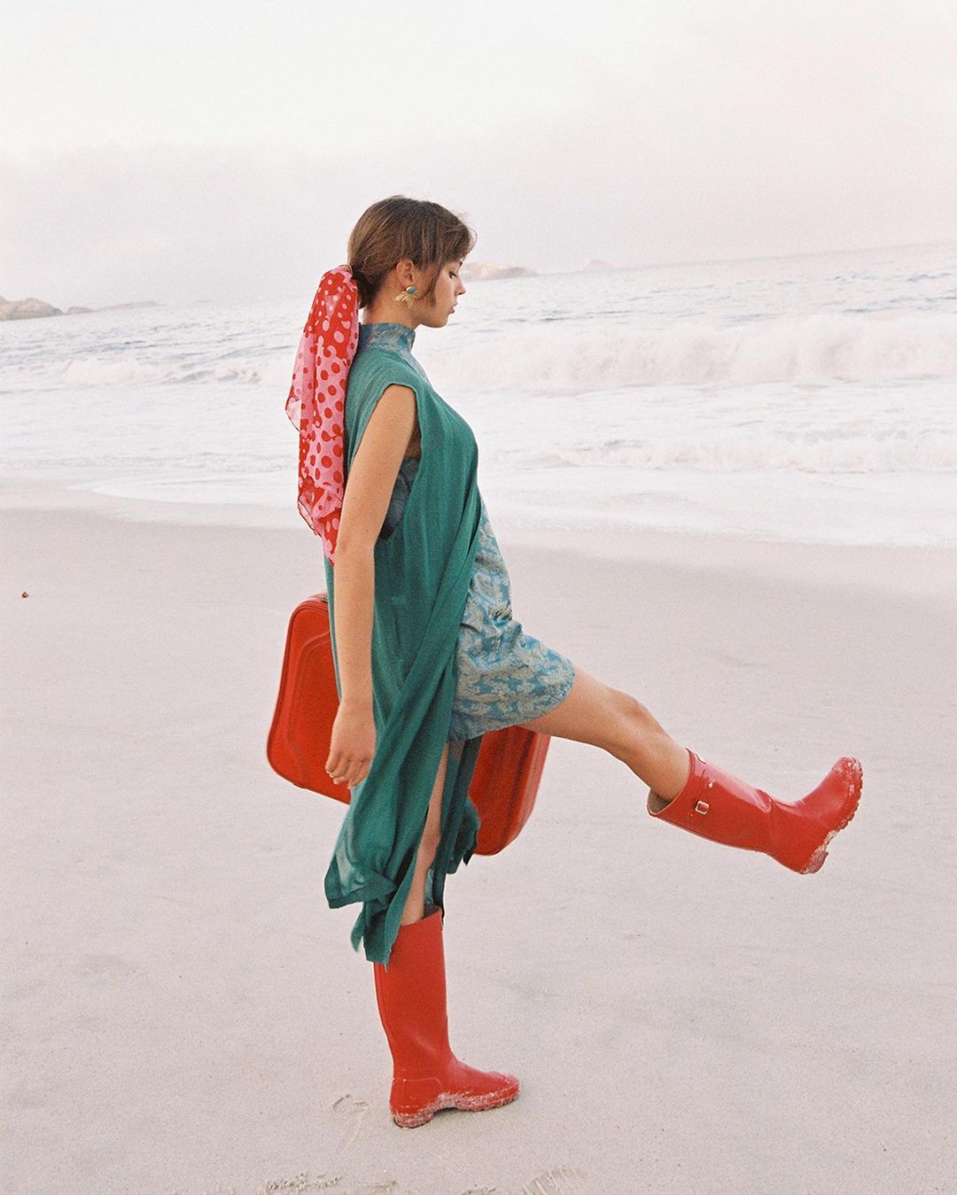 A woman in a teal dress and red boots poses on a sandy beach with ocean waves in the background, holding a red suitcase.