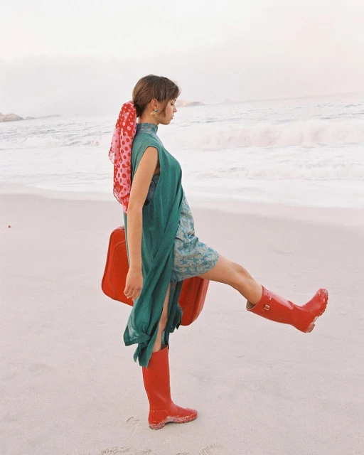 A woman in a teal dress and red boots poses on a sandy beach with ocean waves in the background, holding a red suitcase.