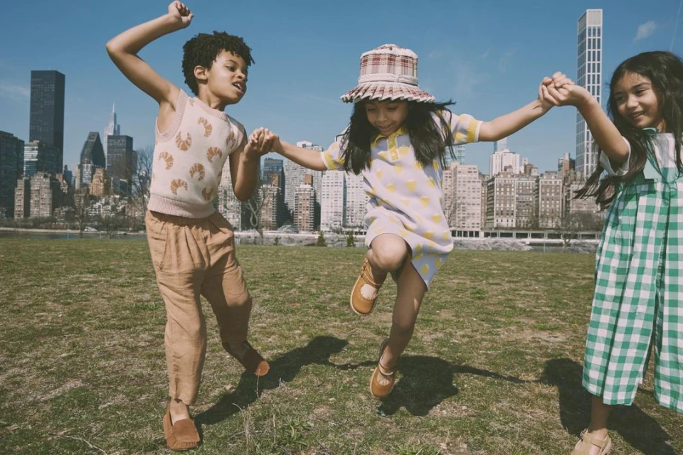 Three children joyfully holding hands in a grassy park with a cityscape backdrop, wearing fashionable clothing, and enjoying a sunny day.