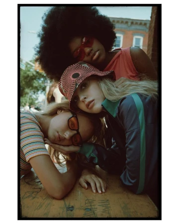 Three young women pose closely together outdoors. One wears a colorful bucket hat, another has an Afro, and all have 1970s style.