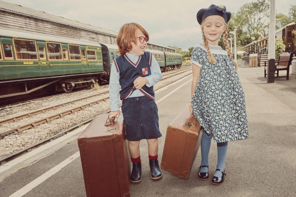 A smiling boy with glasses and a girl with pigtails stand on a train platform with suitcases, dressed in vintage-style children's clothing.
