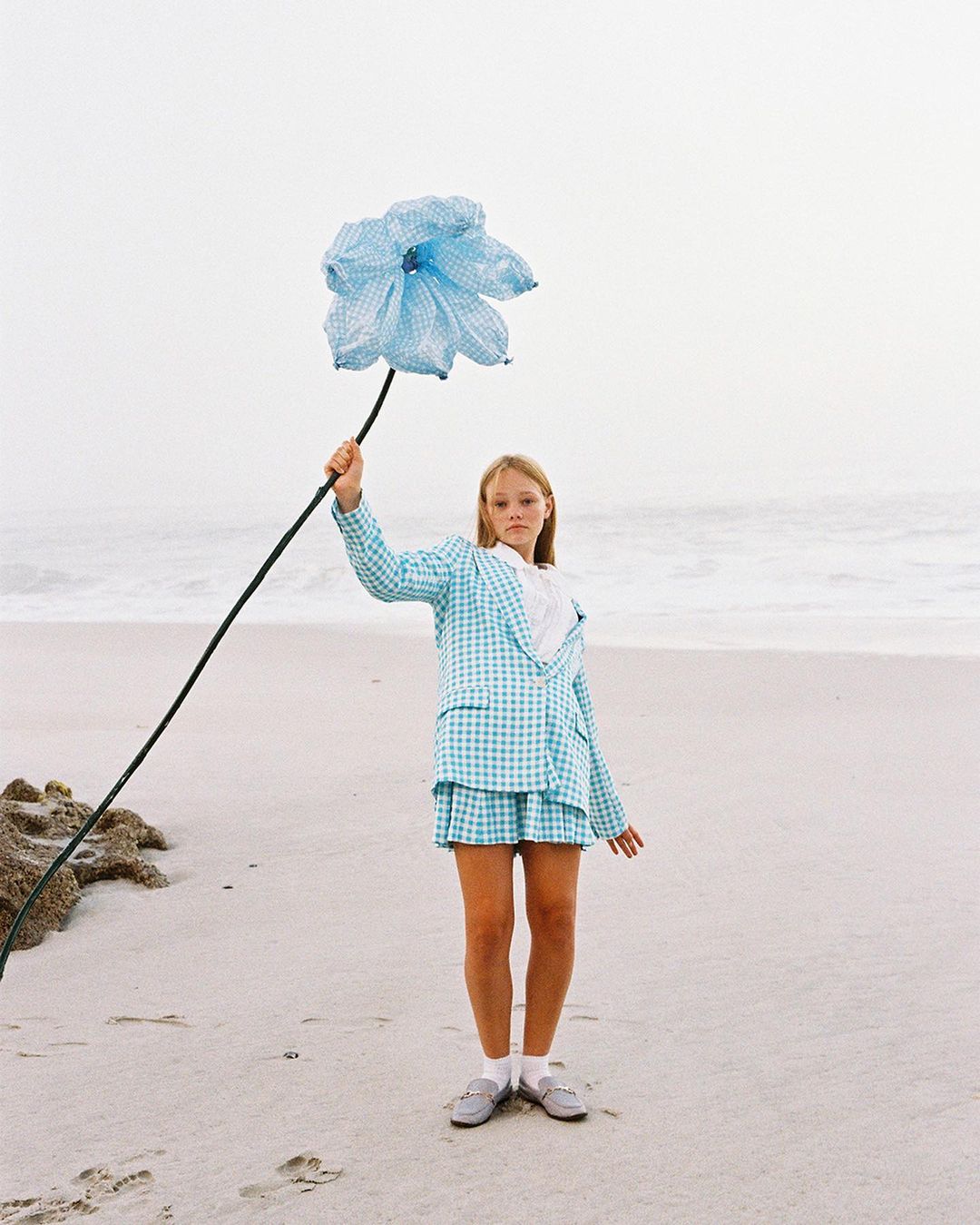 A fair-skinned child in a checkered blue suit stands on a beach holding an oversized blue flower against a backdrop of the ocean and sky.