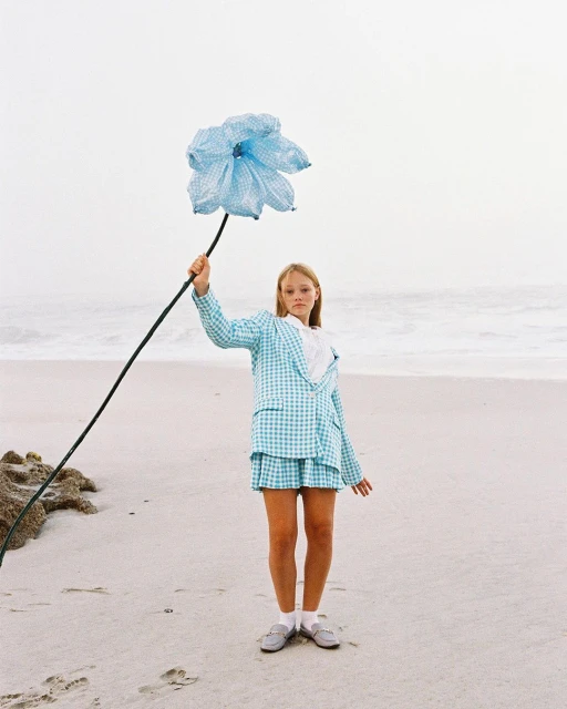 A fair-skinned child in a checkered blue suit stands on a beach holding an oversized blue flower against a backdrop of the ocean and sky.