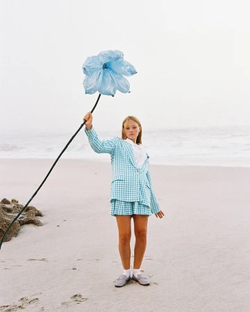 A fair-skinned child in a checkered blue suit stands on a beach holding an oversized blue flower against a backdrop of the ocean and sky.