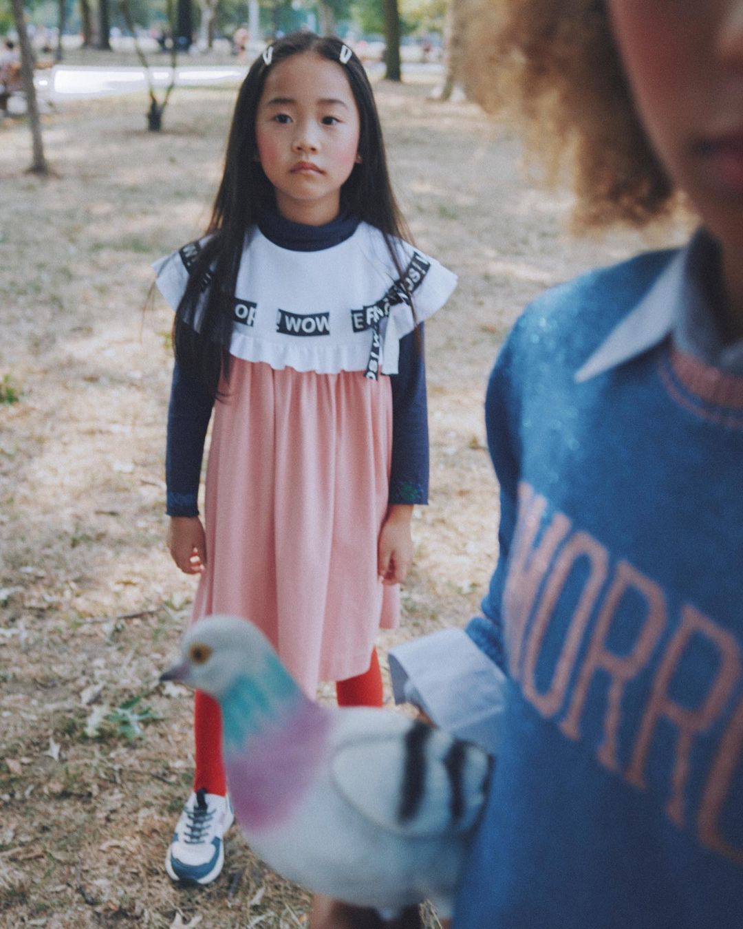 Young girl in a pink dress with word details stands in a park-like setting with another child and a pigeon in the foreground.