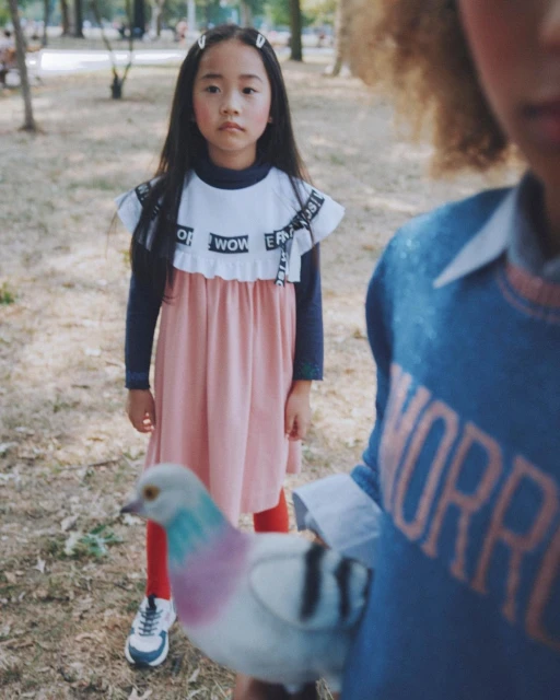 Young girl in a pink dress with word details stands in a park-like setting with another child and a pigeon in the foreground.