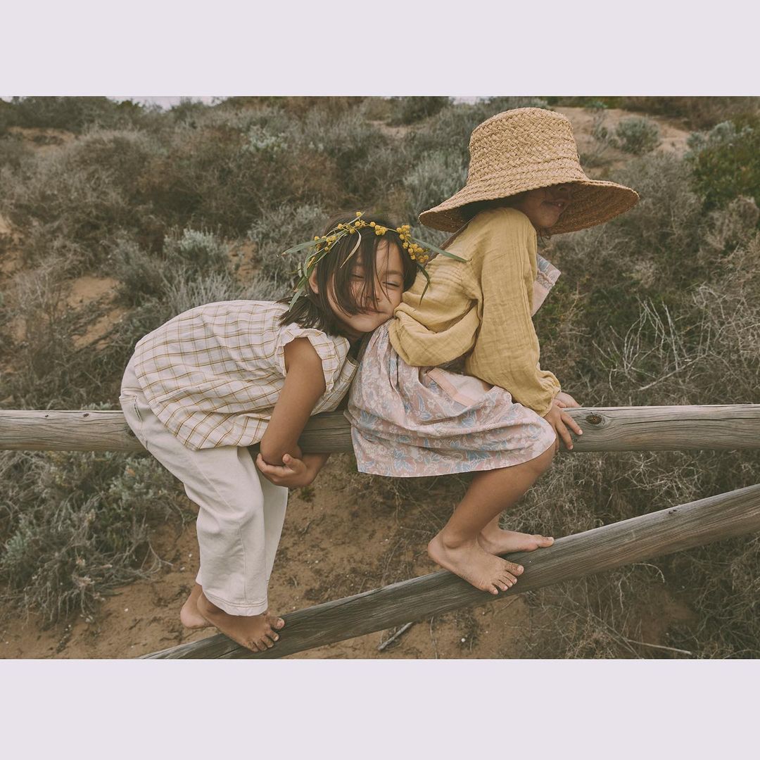 Two young girls in vintage clothing sit on a wooden fence outdoors amidst sandy ground and sparse vegetation, creating a nostalgic scene.