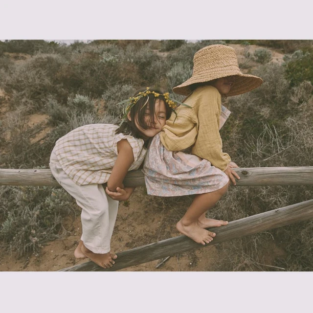 Two young girls in vintage clothing sit on a wooden fence outdoors amidst sandy ground and sparse vegetation, creating a nostalgic scene.