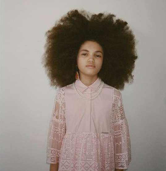 Portrait of a young person with a large Afro hairstyle wearing a pink lace dress and a double-strand pearl necklace against a white background.