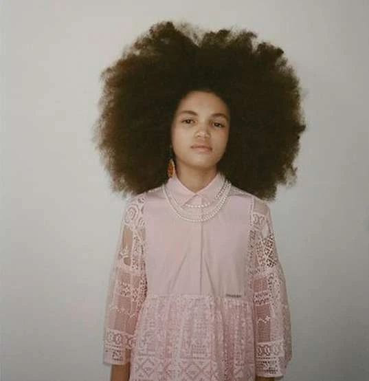 Portrait of a young person with a large Afro hairstyle wearing a pink lace dress and a double-strand pearl necklace against a white background.