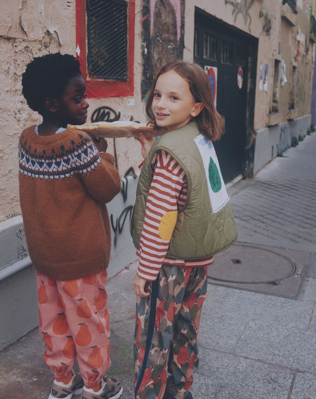 Two children, one Black and one white, carrying a baguette on a city street; dressed in a sweater and vest with printed pants.