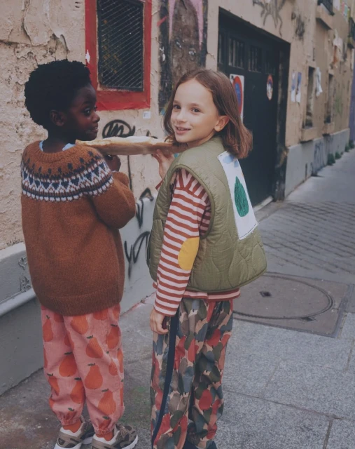Two children, one Black and one white, carrying a baguette on a city street; dressed in a sweater and vest with printed pants.