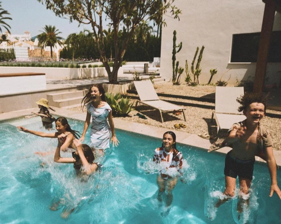 Group of children playing and splashing in a turquoise swimming pool with clear water, surrounded by a villa, deck chairs, and greenery.