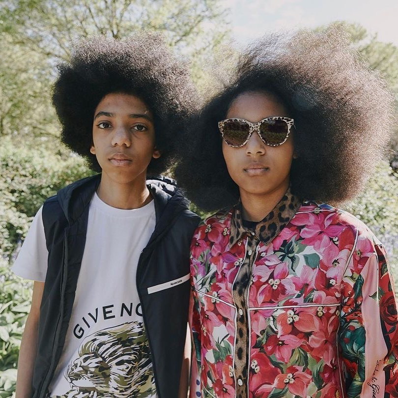 Two young people stand outdoors among trees; the female wears sunglasses and a floral shirt, while the male wears a t-shirt and vest.