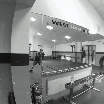 Monochromatic gym shot with an adult male boxer punching a bag, surrounded by equipment and weights, with 'WEST HAM ABC' displayed.