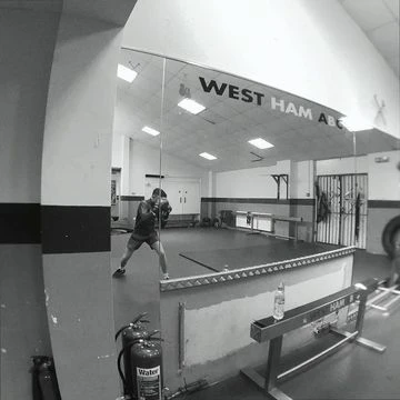 Monochromatic gym shot with an adult male boxer punching a bag, surrounded by equipment and weights, with 'WEST HAM ABC' displayed.