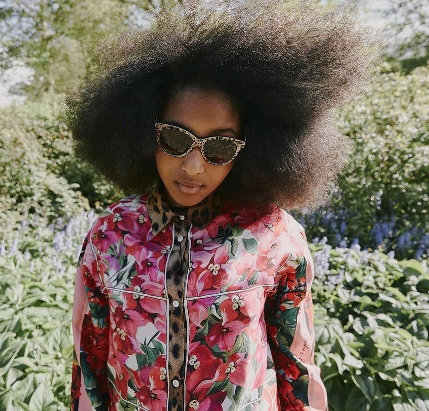 Woman with large afro wears floral top and leopard sunglasses, standing outdoors in a garden setting with diffused light.
