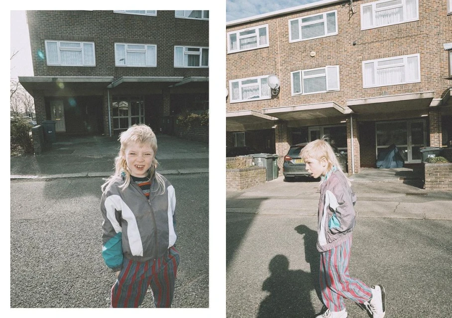 Youngster with mullet hairstyle and striped tracksuit poses outdoors near brick apartments on sunny day, creating a retro-urban aesthetic.