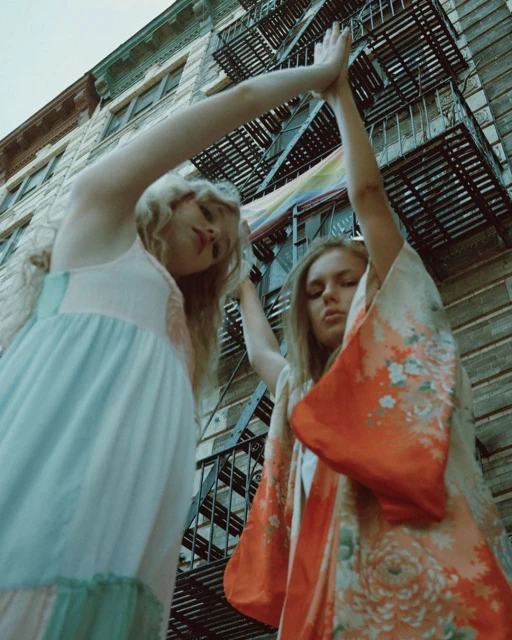 Two women stand under a fire escape of a building, one in a gown and the other in a floral robe, with arms raised overhead in a city.