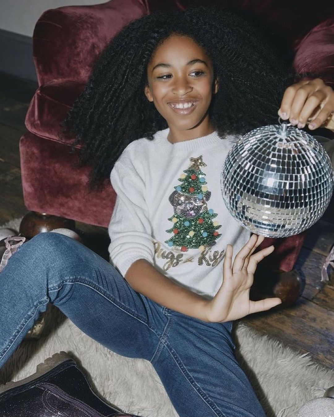 Young girl smiles, holding a disco ball while sitting next to a red chair wearing a Christmas tree-decorated sweater indoors.