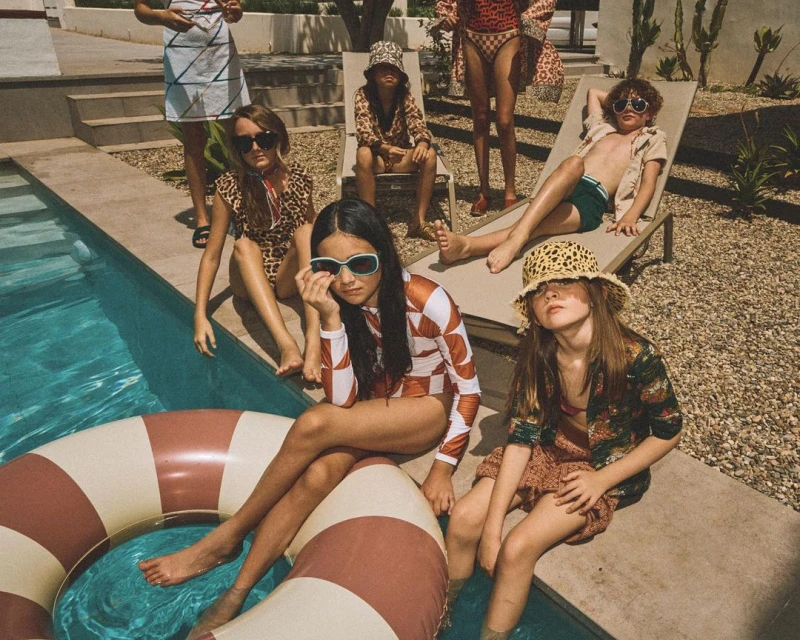 Group of children in swimwear and sunglasses sit and lie by a swimming pool with a float in front of a desert backdrop on a sunny day.