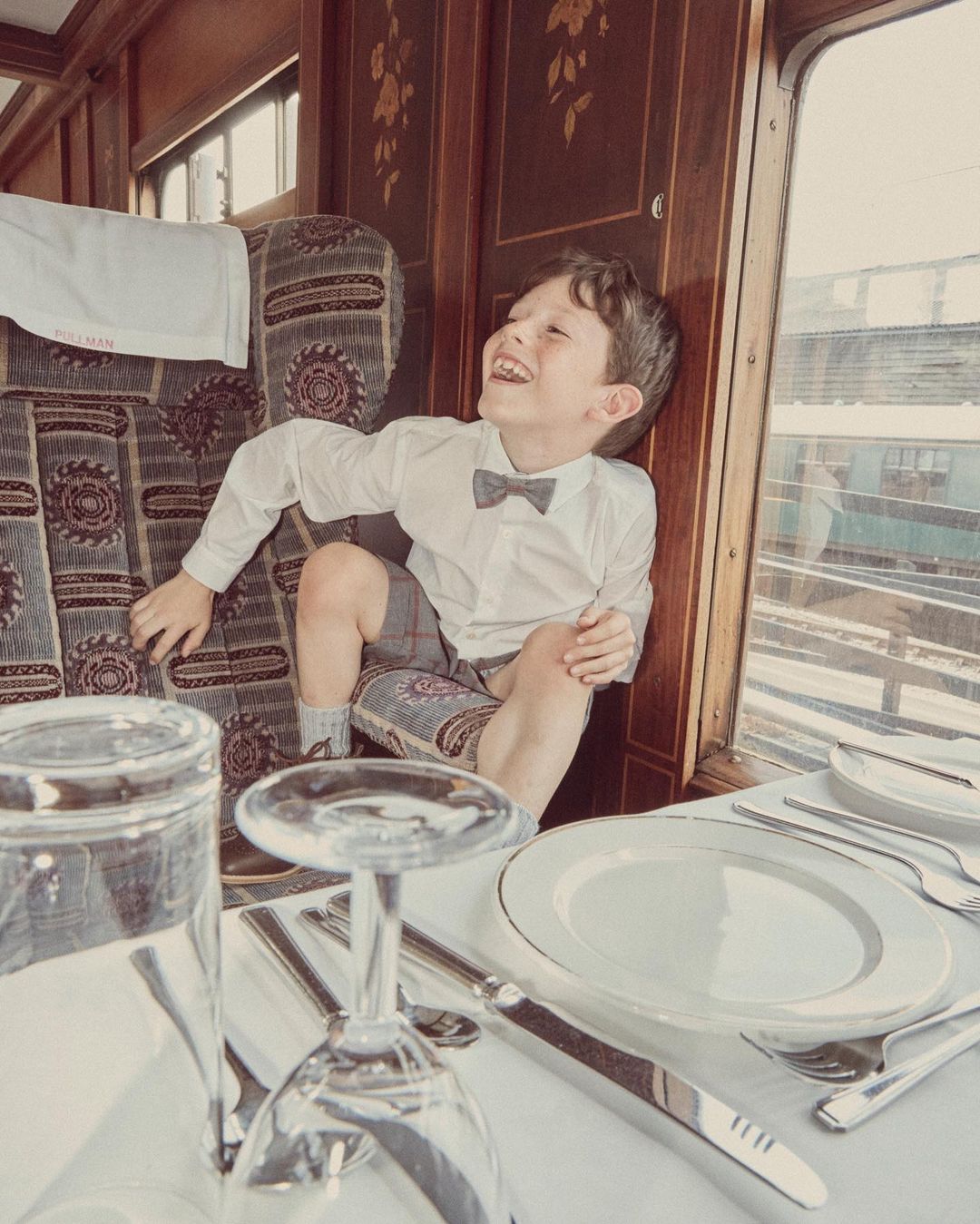 Boy in formal wear laughs inside a vintage train car with set dining table, glasses, silverware, and white table cloth.