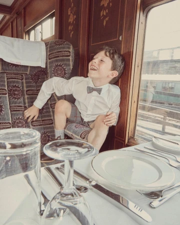 Boy in formal wear laughs inside a vintage train car with set dining table, glasses, silverware, and white table cloth.