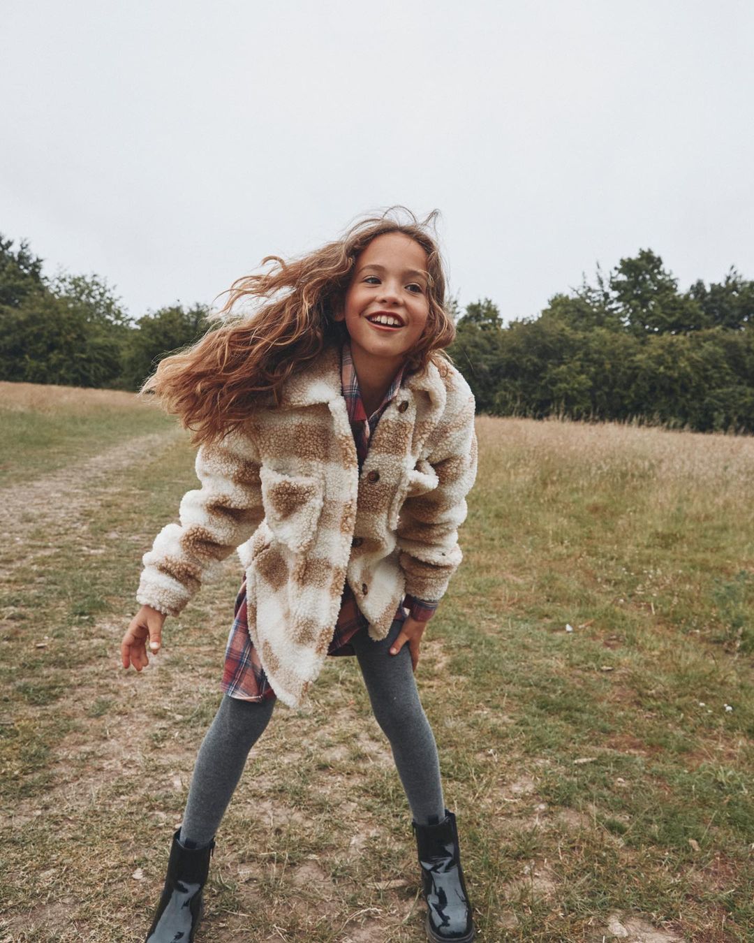 A young girl with curly brown hair smiles outdoors in a plaid sherpa jacket, dress, leggings, and black boots against a nature background.