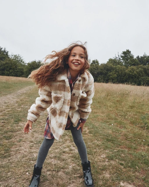 A young girl with curly brown hair smiles outdoors in a plaid sherpa jacket, dress, leggings, and black boots against a nature background.