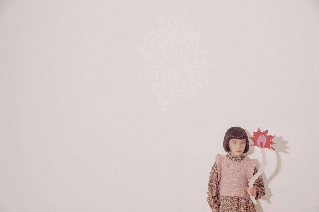 Young girl with short brown hair wearing a floral dress and pink sweater vest holding a flower against a plain white background.