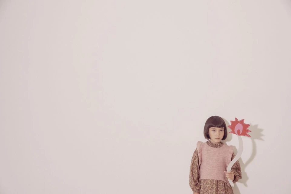 Young girl with short brown hair wearing a floral dress and pink sweater vest holding a flower against a plain white background.