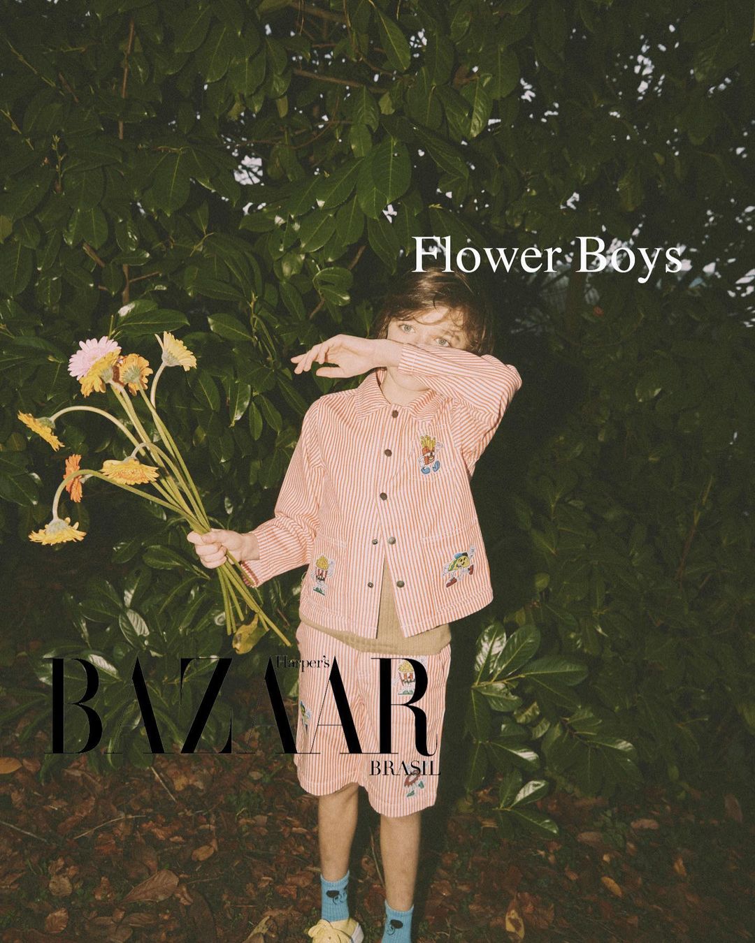 Boy in striped suit with flowers stands in a leafy outdoor setting, fashion editorial for Bazaar Brasil, natural light.